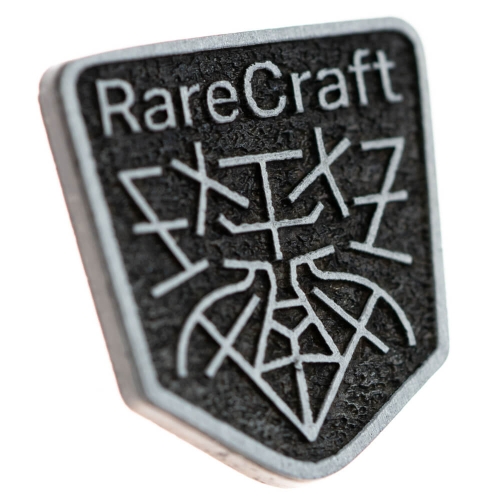 Metalowy emblemat w kształcie heksagonu z napisem RareCraft na jasnym tle. Widoczne geometryczne wzory w czarno-srebrnej kolorystyce. Ujęcie z bliska, pod kątem, eksponujące strukturę i detale zdobienia.