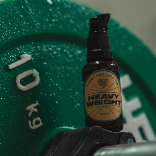 Olejek do brody HEAVYWEIGHT 30 ml w brązowej butelce z czarną pompką na czarnej podstawie, z zielonym ciężarkiem 10 kg w tle. Etykieta z napisami BEARD AND FACE OIL i HEAVY WEIGHT na złotym tle.