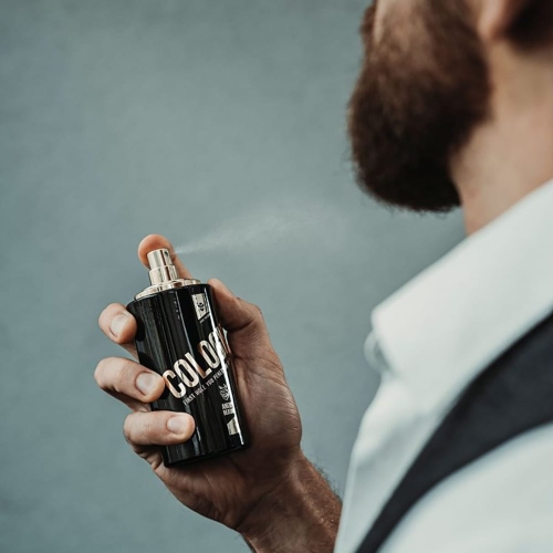 Woda kolońska Angry Beards Jack Saloon w czarnej, prostokątnej butelce, trzymana w dłoni mężczyzny, z rozpylającą się mgiełką na tle szarej ściany, obok napis COLOR.