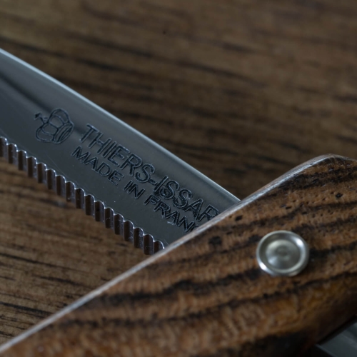 Brzytwa Thiers Issard ze stalowym ostrzem z napisem THIERS-ISSARD MADE IN FRANCE. Rączka z drewna bocote o unikalnym wzorze. Widok zbliżenia na detal, na drewnianym tle z metalowym elementem mocującym.