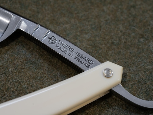 Brzytwa do golenia Thiers Issard Le Dandy White 5/8, z białą rączką i stalowym ostrzem, widoczna z boku. Na ostrzu umieszczony jest napis THIERS: ISSARD MADE IN FRANCE. Tło w jednolitym zielonym kolorze, co podkreśla elegancję produktu.