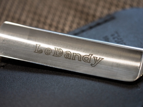 Metalowy klips do krawata z wyraźnym logo LeDandy, leżący na czarnej skórze, w zbliżeniu, z gładką, błyszczącą powierzchnią.