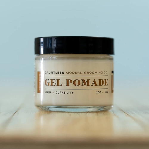 Pomada do włosów w przezroczystym słoiku o czarnej zakrętce, z widocznym napisem GEL POMADE w odcieniu brązu. Produkt leży na drewnianym blacie, pod kątem, ukazując pojemność 56 g. Tło w jasnym kolorze.