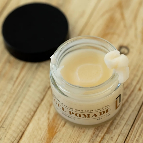 Pomada do włosów w przezroczystym słoiczku ze szklaną pokrywką i czarnym wieczkiem. Biała konsystencja, napis GEL POMADE na etykiecie. Produkt leży na drewnianej powierzchni, widok z góry.