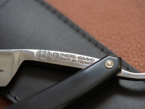 Brzytwa Thiers Issard z wąskim lustrzanym ostrzem i stalowym napisem „THIERS-ISSARD MADE IN FRANCE”, czarną teksturowaną rączką, na czarnym skórzanym tle, podkreślającym jej elegancję i wysoką jakość.