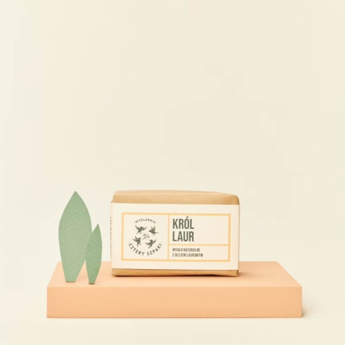 Naturalne mydło Król Laur na pomarańczowej podstawce, w minimalistycznym stylu. Opakowanie z brązowym papierem, z czytelnym napisem KRÓL LAUR oraz logo Mydlarni Cztery Szpaki. W tle widoczne stylizowane zielone liście na jasnym tle.