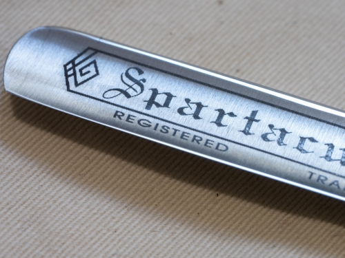 Brzytwa Thiers Issard Spartacus 3/8 z wygrawerowanym logo „Spartacus” oraz napisem „REGISTERED”, ukazana z bliska na gładkiej, metalowej powierzchni. W tle widoczna struktura tkaniny w neutralnym kolorze.