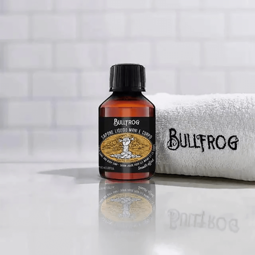 Mydło w płynie Bullfrog w butelce 100 ml, z czarną nakrętka i etykietą z czytelnym logo i ilustracją dłoni. Butelka stoi na gładkiej, połyskliwej powierzchni obok białego ręcznika z haftem. Tło w odcieniach szarości, zapewniające kontrast dla produktu.