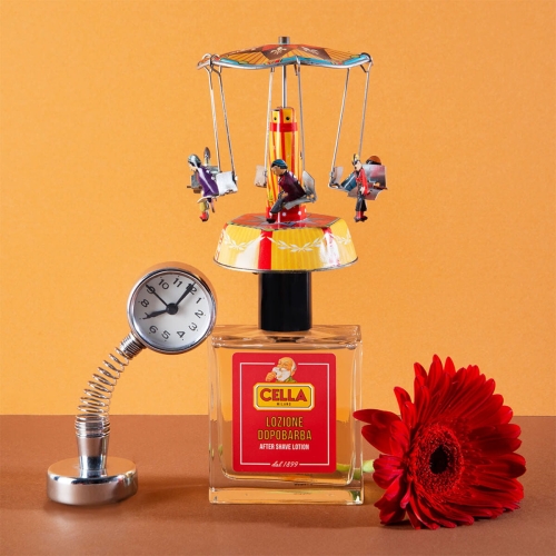 Woda po goleniu Cella Milano 100 ml w eleganckiej szklanej butelce z czerwono-żółtymi detalami i logo, ozdobiona karuzelą z metalowymi figurkami. Obok zegar w chromowanej obudowie na elastycznej podstawie, na pomarańczowym tle z czerwoną gerberą.