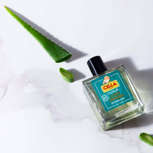 Woda po goleniu Cella Milano BIO Aloe Vera 100 ml w przezroczystej butelce z czarną nakrętką na marmurowym tle, obok liście aloesu i fragmenty miąższu. Etykieta z logo i napisami marki Cella.