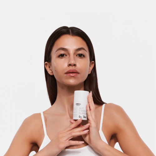 Kobieta prezentuje krem SPF50+ 50 ml w białym opakowaniu z napisem BUT FIRST SUNSCREEN i logo veoli na białym tle. Ma biały top i delikatny makijaż, podkreślając codzienną pielęgnację skóry.