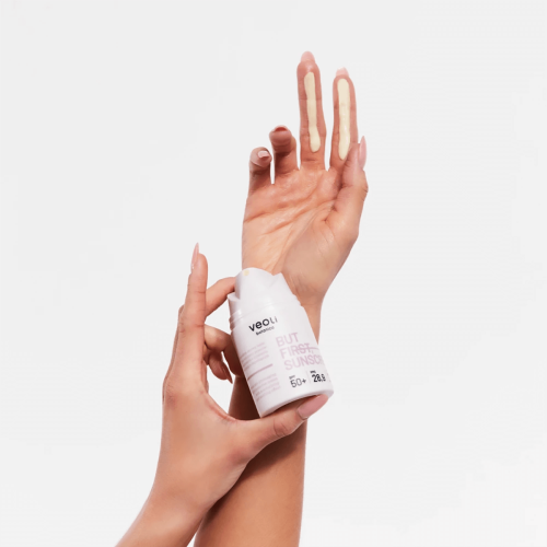 Krem ochronny SPF50+ Veoli Botanica w białej tubce z różowymi napisami, trzymany w dłoni. Palce drugiej ręki pokazują dwie smugi na skórze, ilustrując teksturę i aplikację produktu. Tło jest jednolite, co podkreśla detal.