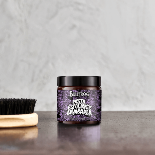 Peeling do brody Bullfrog 100 ml w stalowym słoiku z czarną nakrętką, ozdobionym fioletowo-czarnym wzorem. Leży na ciemnym blacie obok szczotki z czarnym włosiem i drewnianą rączką. Jasne tło podkreśla etykietę z nazwą i opisem.
