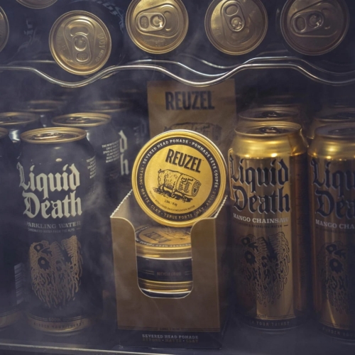 Matowa pomada Reuzel x Liquid Death Severed Head w złotym opakowaniu na półce z złotymi puszkami Liquid Death. Okrągły pojemnik z logo Reuzel na tle ciemnej, zamglonej tekstury.