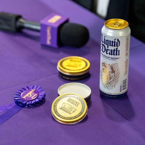 Matowa pomada Reuzel w pojemniku z czarnym dnem i złotą pokrywką, obok zamkniętego słoika i puszki Liquid Death, na fioletowym tle przy mikrofonie. Widoczna purpurowa wstążka z napisem 1st.