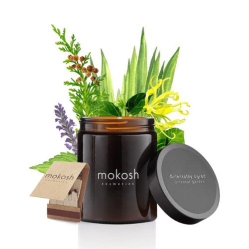 Świeca Mokosh Orientalny Ogród 180 ml w ciemnym szkle z napisem i otwartą pokrywką. W tle zielone liście i kwiaty, podkreślające naturalny aromat. Słoik na jasnym tle.