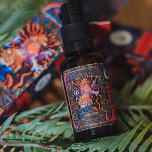 Olejek do brody w butelce 30 ml z czarną pompką, z kolorową etykietą przedstawiającą wschodzące słońce i ornamenty, leżący na zielonym tle iglastych gałęzi. W tle widoczna, ozdobna pudełkowa grafika.