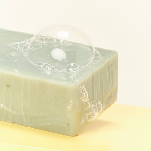 Mydło eukaliptus cytrynowy w jasnooliwkowym kolorze, z widoczną bąbelkową pianą na górze. Produkt leży na jednolitym, jasnym tle. Kąt ujęcia zbliżony na detal mydła, ukazujący jego gładką fakturę oraz naturalne drobinki.