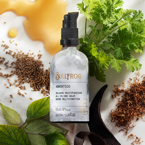 Bullfrog Agnostico to 100 ml balsam w owalnej, przezroczystej butelce z czarną pompką i etykietą w białe, czarne i złote napisy. Leży na tle z liśćmi, ziarnami i miodem, podkreślając naturalne składniki i łatwość użycia.