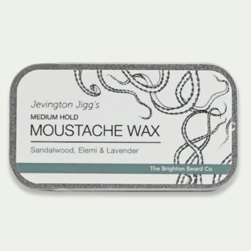 Wosk do wąsów w metalowym opakowaniu z grafiką. Widoczny napis Jevington Jigg's Medium Hold Moustache Wax, składniki: Sandalwood, Elemi & Lavender. Produkt leży na jasnym tle, nadającym się do minimalistycznych aranżacji.
