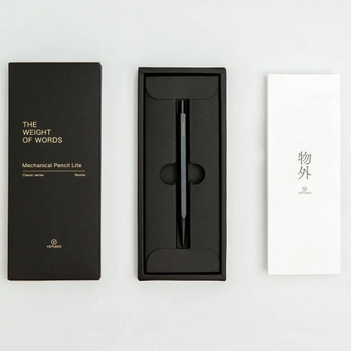 Czarny ołówek mechaniczny YSTUDIO Classic Revolve Lite w eleganckim pudełku z złotą grafiką „The Weight of Words”, plus biały arkusz z logo YSTUDIO, leżący na jasnym tle.