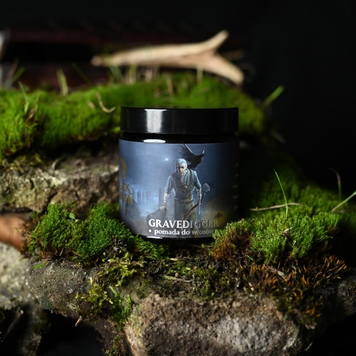 Slickhaven pomada do włosów Gravedigger 60 ml w matowym, czarnym słoiku z kolorową etykietą przedstawiającą postać na tle zielonego mchu. Widoczne napisy GRAVEDIGGER i pomada do włosów. Naturalne tło z elementami przyrody.