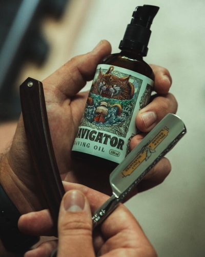 Olej do golenia Navigator 100 ml w brązowej butelce z dozownikiem, etykieta z lwem i napisami NAVIGATOR oraz SHAVING OIL 100 ml. W drugiej ręce klasyczna golarka z metalowym ostrzem i drewnianą rączką na rozmytym tle.