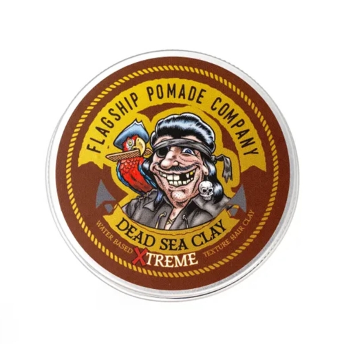 Ciemno-brązowa, okrągła puszka z kolorowym nadrukiem pirata z papugą. Napisy: FLAGSHIP POMADE COMPANY, DEAD SEA CLAY oraz informacja o bazie wodnej. Puszka zamknięta, widoczna frontalnie.