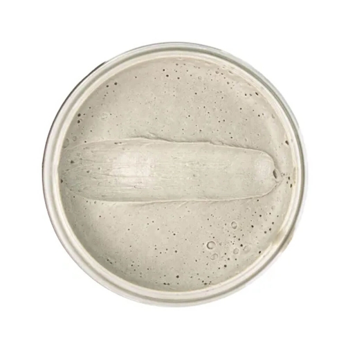 Woda pomada do włosów w pojemniku, widok z góry. Produkt o szarej, gładkiej strukturze, z wyraźnymi bąbelkami powietrza. Kształt okrągły, z widocznym centrum, gdzie widać, że produkt jest zdrapywany. Brzeg pojemnika metaliczny.