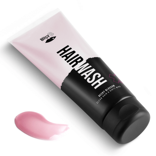 Różowy szampon BECKY BLOSSOM z ciemnoszarym, falistym spodem, napis HAIR WASH w białej czcionce. Butelka stoi lekko z boku obok różowej kropelki szamponu na gładkiej powierzchni.