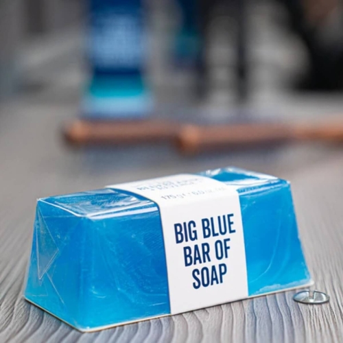 Błękitne mydło w kostce o masie 175 g, z widoczną etykietą z napisem BIG BLUE BAR OF SOAP. Produkt leży na drewnianej powierzchni, w zbliżeniu na detal, ukazując gładką, przezroczystą teksturę.