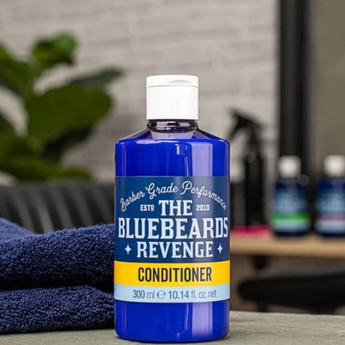 Odżywka do włosów THE BLUEBEARDS REVENGE w błękitnej butelce 300 ml z białą zakrętką, na szarej powierzchni obok niebieskiego ręcznika. W tle zielone liście i inne butelki.
