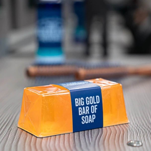 Mydło w formie dużej, złotej kostki (175 g) w pomarańczowym kolorze, z niebieską etykietą z napisem BIG GOLD BAR OF SOAP. Produkt leży na drewnianym blacie, w tło widoczne są nieostre przedmioty. Etykieta jest czytelna i dobrze widoczna.