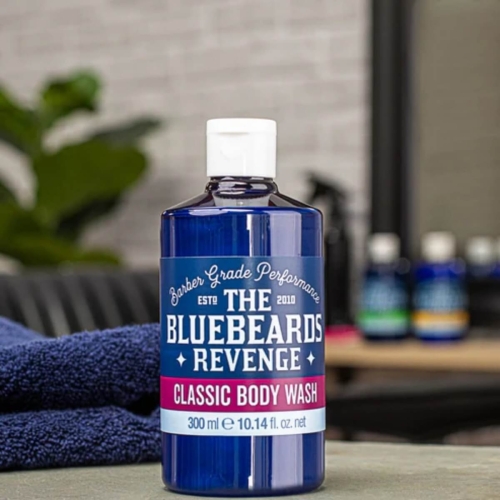 Butelka żelu Bluebeards Revenge 300 ml, błękitna z białą zakrętką, napis CLASSIC BODY WASH i logo. Na żółto-szarym tle z niebieskim ręcznikiem i wielobarwnymi produktami w tle.