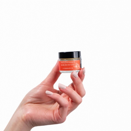 Orzeźwiające serum do ust z pomarańczą w szklanym słoiczku trzymanym w dłoni, z widocznym, pomarańczowym etykietą i czarną pokrywką. Produkt w pozycji pionowej, na jednolitym tle.