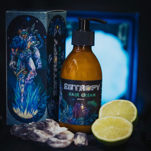 Brązowa butelka 200 ml z czarną pompką i napisem HAIR CREAM na zielono-niebieskiej etykiecie, na ciemnym tle. Obok plasterki limonki i kryształy, w tle kolorowe opakowanie. Widok z boku eksponujący detale.