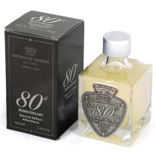 Balsam po goleniu Saponificio Varesino 80th Anniversary w przezroczystej butelce z metalową zakrętką, obok czarnego opakowania z białym nadrukiem i herbem, na jednolitym tle, ujęcie frontalne podkreślające detale.