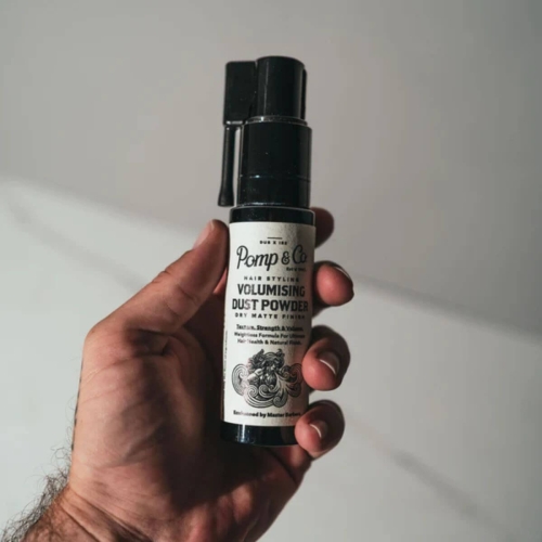 Puder do włosów Pomp&Co. w czarnej butelce z białą etykietą, trzymany w dłoni; widoczny napis „Volumising Dust Powder” oraz logo Pomp&Co. Opakowanie o prostym kształcie z atomizerem, ukazujące matowe wykończenie, na jasnym tle.