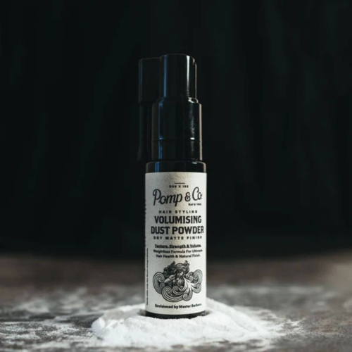 Czarny pojemnik z dozownikiem na puder do włosów o matowym wykończeniu. Biała etykieta z napisem Volumising Dust Powder. Puder rozsypany na szarej powierzchni, pojemnik stoi pionowo na ciemnym tle.