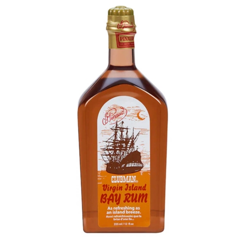 Prostokątna, szklana butelka z przezroczystego szkła z brązowym płynem. Etykieta z białym tłem, ilustracją statku i pomarańczowym napisem CLUBMAN Virgin Island BAY RUM. Złota nakrętka z logo PINAUD. Zdjęcie frontalne na jednolitym tle.