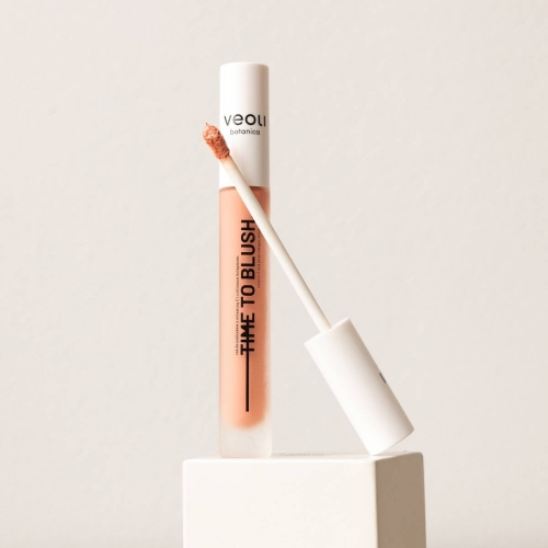 Róż do policzków Veoli Botanica TIME TO BLUSH w odcieniu brzoskwiniowym, w przezroczystym opakowaniu z czarną etykietą. Aplikator w formie gąbeczki, uniesiony w pozycji pionowej. Produkt ustawiony na białym dysku, z tłem w delikatnym beżu.