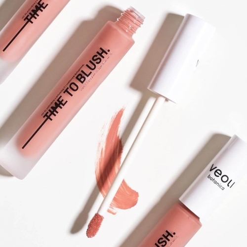 Róż do policzków Veoli Botanica TIME TO BLUSH w odcieniu brudnego różu, zamknięty w przezroczystej, matowej butelce z białą nakrętką. W zestawie znajduje się aplikator z miękkim, włosianym końcem, obok widać smużkę produktu na białym tle.