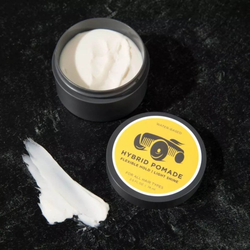 Elastyczna pomada do włosów o lekkim połysku w ciemnym pojemniku. Biała konsystencja na łyżeczce obok. Etykieta z napisem HYBRID POMADE i ikoną samochodu na żółtym tle. Czarne tło podkreśla teksturę pomady.