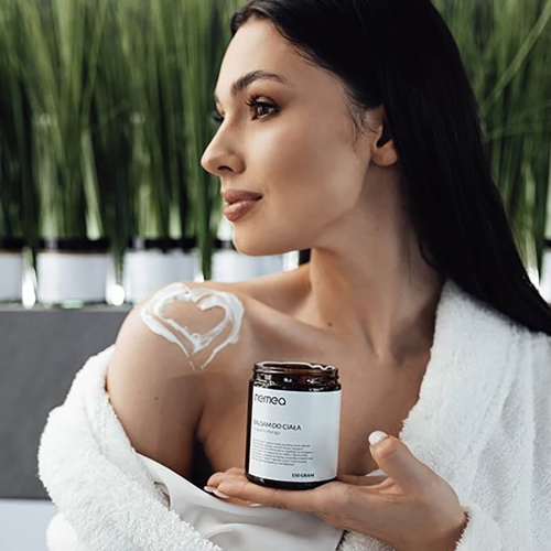 Antycellulitowy balsam do ciała Nemea o pojemności 150 g w ciemnym, szklanym słoiczku, trzymany przez kobietę w białym, puszystym ręczniku. Na ramieniu modelki widoczna jest aplikacja balsamu w kształcie serca. Tło z zielonymi roślinami w doniczkach.