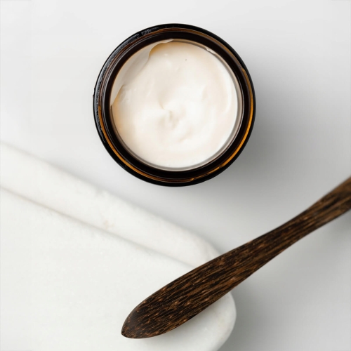 Antycellulitowy balsam do ciała w opakowaniu słoika o ciemnym kolorze, z białą, gładką konsystencją. Widać drewnianą szpatułkę obok, na jasnym tle. Słoik jest zakręcony, widok z góry.