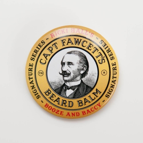 Okrągłe złote pudełko balsamu do brody Captain Fawcett's z ilustracją mężczyzny w eleganckim ubraniu i wąsami, na białym tle, z napisami: Captain Fawcett's Beard Balm i Booze and Baccy, ujęcie frontalne.
