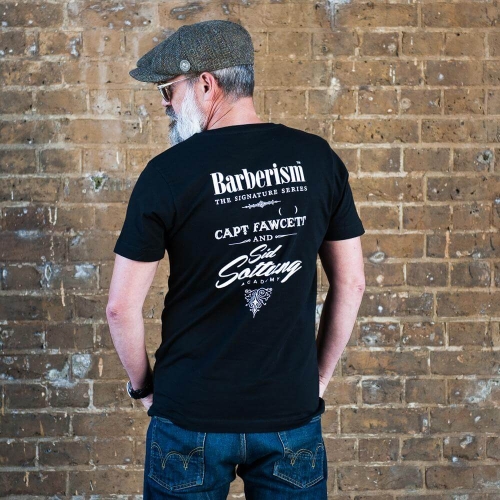 Czarny t-shirt z logo Barberism i białym nadrukiem The Signature Series, Capt Fawcett oraz Sid Sottung Academy na plecach. Mężczyzna w czapce, siwej brodzie i granatowych jeansach stoi przy ceglanej ścianie.