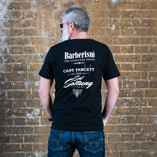 Czarny t-shirt z logo Barberism i napisem CAPT FAWCETT AND Sid Sottung Academy na plecach, noszony przez siwowłosego mężczyznę w okularach, stojącego przy ceglanej ścianie. Luźno dopasowany, z krótkim rękawem, jasne dżinsy.