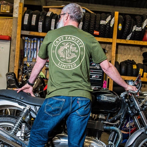 Mężczyzna z białą brodą stoi tyłem w oliwkowej koszulce z napisem CAPT FAWCETT LIMITED i niebieskich dżinsach, trzymając rękę na siedzeniu motocykla Triumph w warsztacie z oponami i narzędziami.