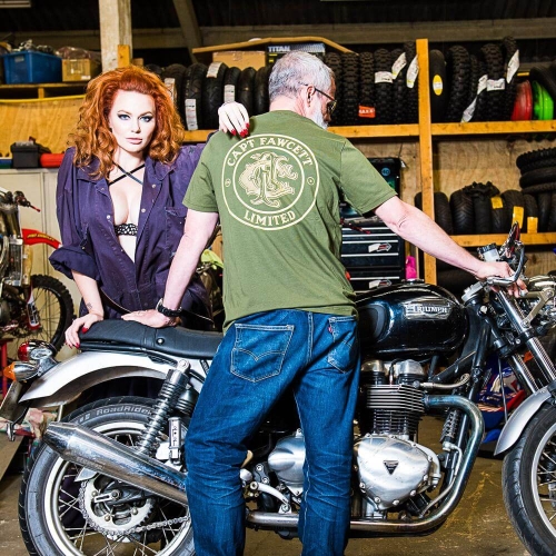 Kobieta w rozpiętej ciemnej koszuli i czarnym biustonoszu stoi obok mężczyzny w zielonej koszulce CAPT FAWCETT LIMITED przy motocyklu Triumph, w warsztacie z oponami i narzędziami na półkach.